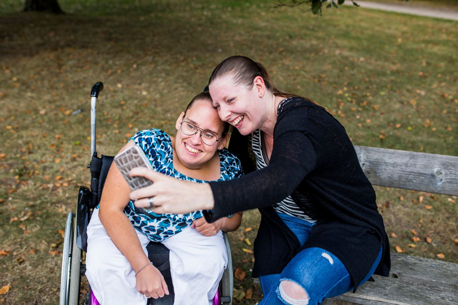Two friends smiling and taking a selfie in a park, one using a wheelchair